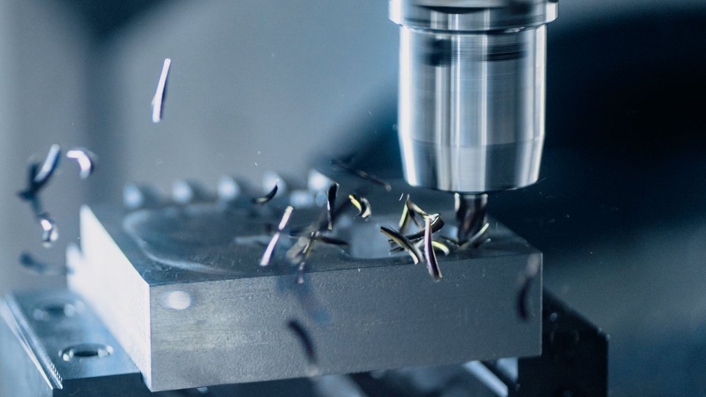 ดอกเอ็นมิลจับในหัวจับของเครื่อง CNC กำลังกัดก้อนเหล็กบนปากกาจับงาน เศษเหล็กม้วนเป็นเกลียวกระเด็นออกจากจุดตัด